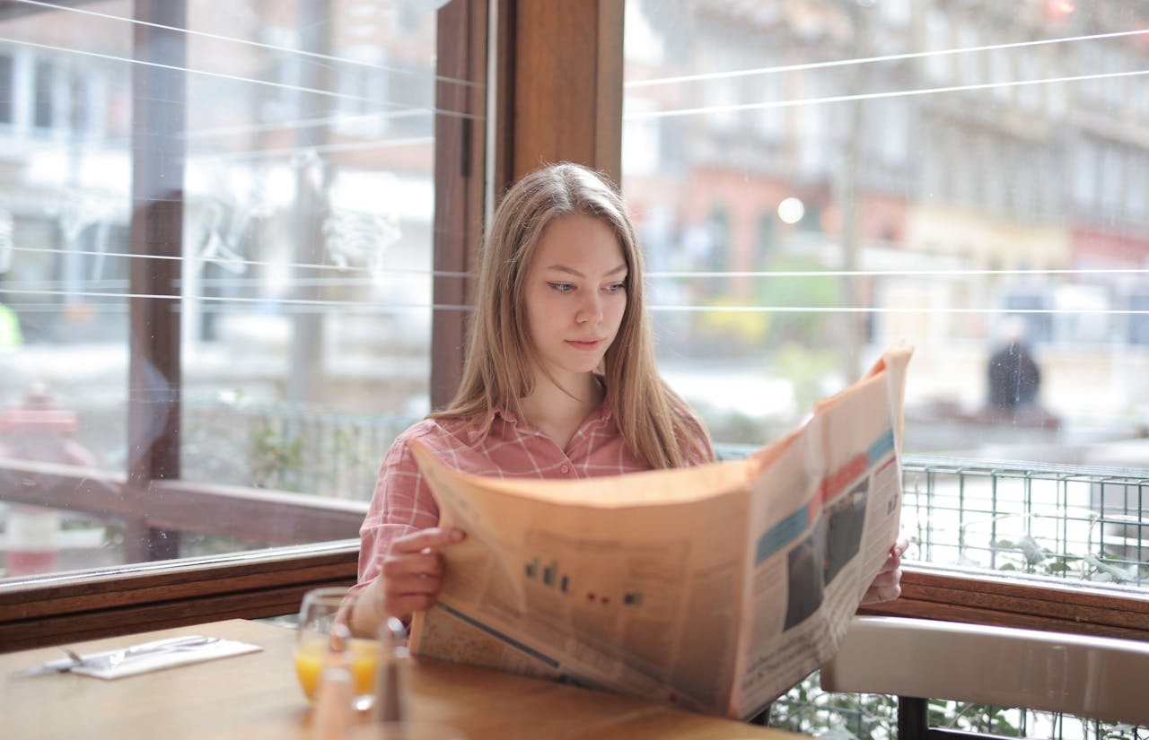 A young woman enjoying a newspaper at a cafe table, immersed in reading.